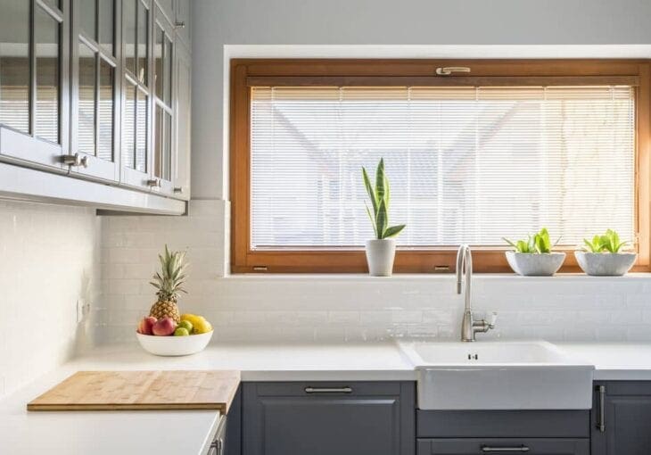 Modern white kitchen with wooden-framed window featuring a light-filtering kitchen blind and small potted plants on the sill